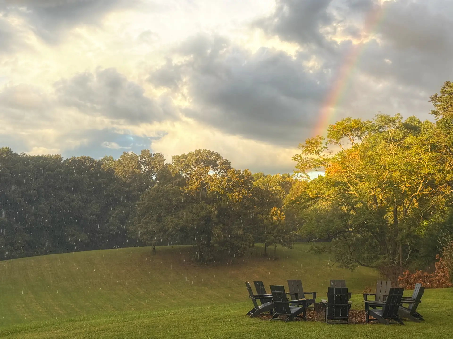 Rain still falling as a rainbow comes down over the fire pit circle, storm light on the trees